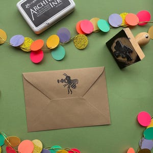 May include: A brown envelope with a black bee illustration holding a trumpet. The envelope is surrounded by a colorful garland of circles with gold glitter.