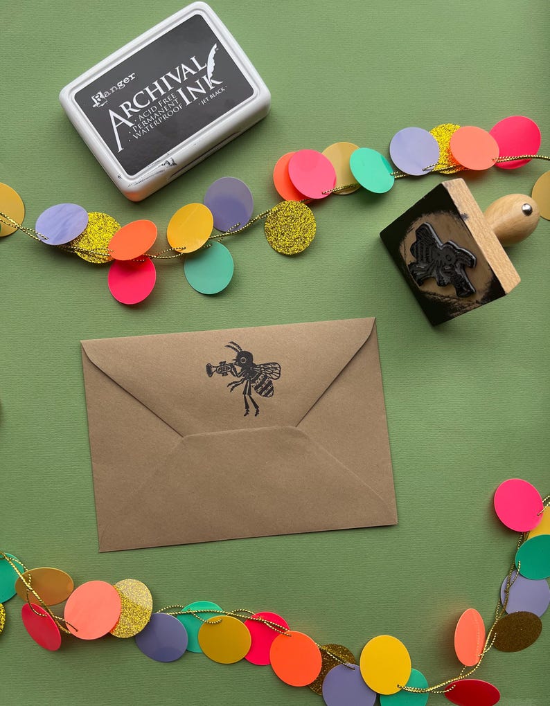 May include: A brown envelope with a black bee illustration holding a trumpet. The envelope is surrounded by a colorful garland of circles with gold glitter.