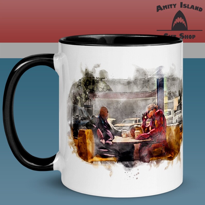 May include: White ceramic coffee mug with a black rim and handle. The mug features a watercolor illustration of two men sitting at a table in a diner. The men are wearing suits and one is holding a cup of coffee. The text "Amity Island Gift Shop" is visible in the upper right corner of the mug.