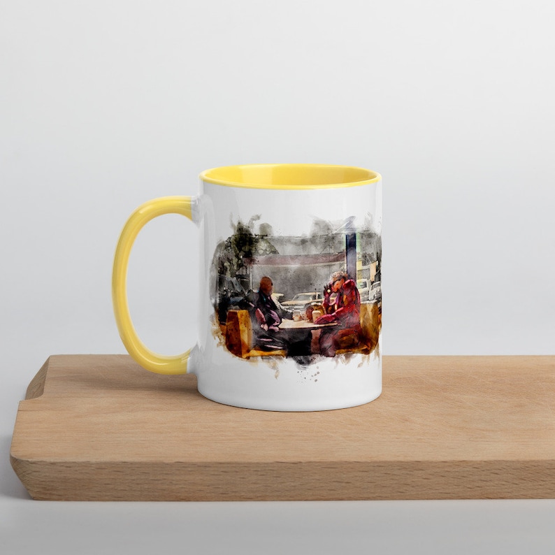 May include: A white ceramic mug with a yellow rim, featuring a watercolor illustration of two people sitting at a table in a diner. The illustration is in shades of brown, red, and yellow.