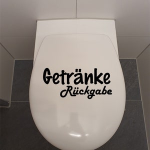 Könnte beinhalten: Ein weißer Toilettensitz mit einem schwarzen Vinyl-Aufkleber, auf dem "Getränke Rückgabe" steht.