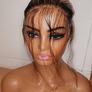 May include: A mannequin head with a brown wig, dark eyebrows, and pink lipstick. The mannequin has a tan complexion and is positioned against a white wall and a wooden floor.