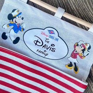 May include: A white canvas bag with red and white stripes. The bag has a wooden dowel at the top for hanging. The bag has a Disney Cruise Line logo and the text "The Davis Family". Mickey Mouse and Minnie Mouse are depicted on the bag in their captain's uniforms.