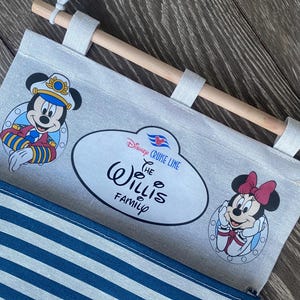 May include: A white canvas wall hanging with blue and white stripes. The top of the hanging has a wooden dowel rod with a rope attached. The hanging features a Mickey Mouse and Minnie Mouse design with the text "Disney Cruise Line The Willis Family".