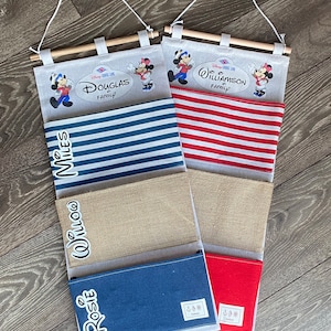 May include: Two white canvas wall organisers with red and blue striped pockets. The organisers feature a Disney Cruise Line logo and Mickey Mouse characters. The top pocket of each organiser has a personalised name, "Douglas Family" and "Williamson Family".