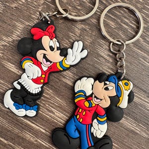 May include: Two Disney character keyrings featuring Minnie Mouse and Mickey Mouse. Minnie Mouse is wearing a red jacket, black skirt, and a red bow. Mickey Mouse is wearing a red jacket and blue trousers. Both keyrings have silver key rings.