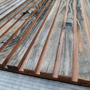 May include: Close-up of a wooden wall panel made of thin, weathered planks arranged in a vertical pattern. The planks are a mix of light brown and gray, with a natural wood grain.