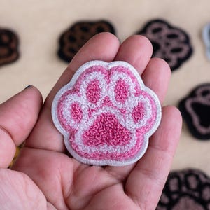 Könnte beinhalten: Ein rosa-weiß gestickter Pfotenabdruck-Patch, der in einer Hand gehalten wird, mit anderen Pfotenabdruck-Patches in verschiedenen Farben im Hintergrund verstreut. Die Patches sind wahrscheinlich für Kleidung oder Accessoires.