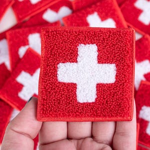 May include: A collection of red square patches with a white cross design, resembling the Swiss flag. The patches are made of a textured fabric and are scattered on a white surface. One patch is held in a hand.