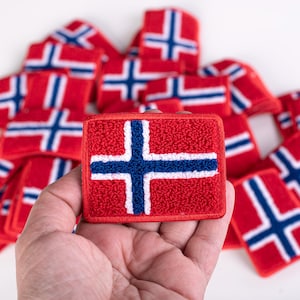 Könnte beinhalten: Ein rechteckiger bestickter Aufnäher mit der norwegischen Flagge, bestehend aus rotem Hintergrund, blauem Kreuz und weißen Rändern. Der Aufnäher hat eine texturierte Oberfläche aus Schlingen und wird vor einem Stapel gleicher Aufnäher gehalten.