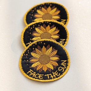 Könnte beinhalten: Drei schwarze Stoffpatches mit gelben gestickten Sonnenblumen. Der untere Patch hat den Text "Face the Sun" in Gelb gestickt.