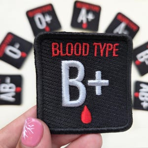 Könnte beinhalten: Ein quadratischer, schwarzer, bestickter Aufnäher mit dem roten Text BLOOD TYPE über einem großen weißen B+ Symbol und einem kleinen roten Blutstropfen-Symbol. Mehrere weitere ähnliche Aufnäher mit verschiedenen Blutgruppen sind im Hintergrund zu sehen.