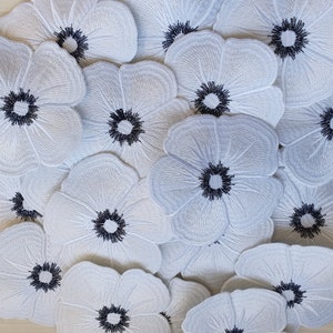 Pode incluir: Uma coleção de patches de flores de feltro branco com centros bordados em preto. Os patches estão dispostos em uma pilha, mostrando o detalhe do bordado.