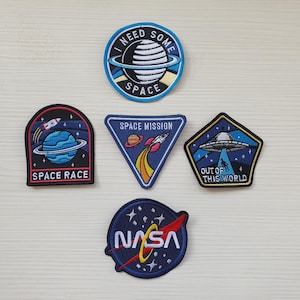 May include: A collection of embroidered space-themed patches. Designs include a planet with the text "I NEED SOME SPACE", a rocket ship, a UFO, and the NASA logo. Patches are in various shapes and colors.