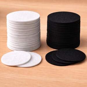 Op de afbeelding: Twee stapels ronde onderzetters, één witte en één zwarte. De onderzetters zijn gemaakt van vilt. Een paar onderzetters liggen plat voor de stapels.