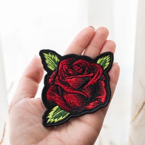 Op de afbeelding: Een rood geborduurd rozenpatch met groene bladeren. Het patch bevindt zich op een hand.