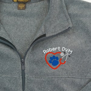 May include: Gray fleece zip-up jacket with the text "Robert, DVM" embroidered in white, a red heart, a blue paw print, and a stethoscope. The jacket has a black zipper and a collar. The brand name "Harriton" is visible on the upper left.
