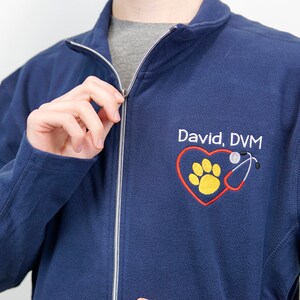 Puede incluir: Una chaqueta de forro polar azul marino con cierre de cremallera. La chaqueta tiene un logotipo bordado con un corazón rojo, una huella de pata amarilla y un estetoscopio rojo. El texto "David, DVM" está bordado debajo del logotipo.