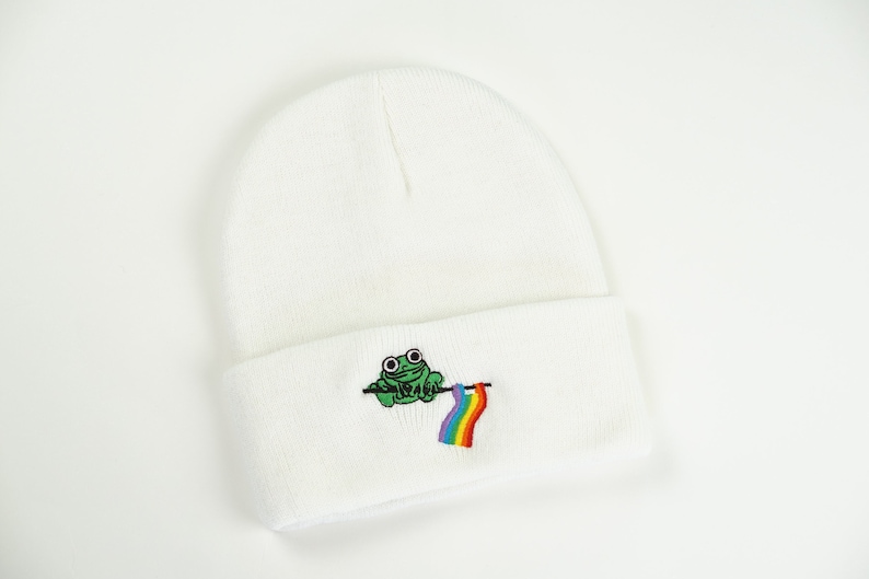 May include: White beanie hat with an embroidered green frog holding a rainbow flag. The hat is made of a soft knit material. The frog has large, round eyes. The rainbow flag has stripes of red, orange, yellow, green, blue, and purple.