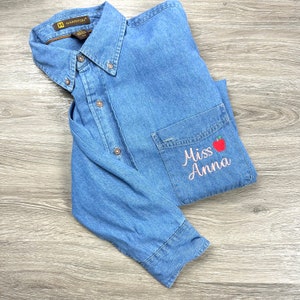 Pode incluir: Uma camisa de ganga azul com um bolso que tem o texto "Miss Anna" bordado em fio rosa. Uma maçã vermelha com um caule verde está bordada acima do texto.