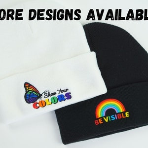 May include: Two beanies are shown, one white and one black. The white beanie has a colourful butterfly and the text "Show Your COLORS." The black beanie features a rainbow and the text "BE VISIBLE." The words "MORE DESIGNS AVAILABLE" are at the top.