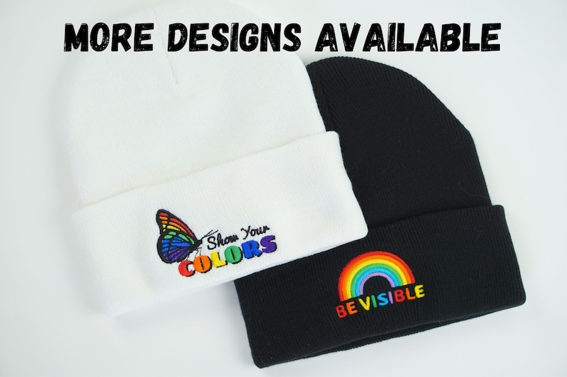 May include: Two beanies are shown, one white and one black. The white beanie has a colourful butterfly and the text "Show Your COLORS." The black beanie features a rainbow and the text "BE VISIBLE." The words "MORE DESIGNS AVAILABLE" are at the top.
