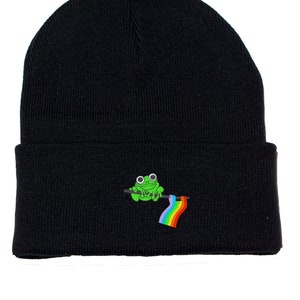 May include: Black beanie hat with an embroidered design of a green frog holding a rainbow flag. The hat is made from a knitted fabric. The frog has large, round eyes and is perched on a dark green branch.