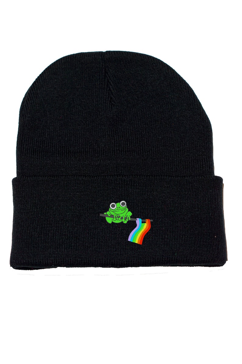 May include: Black beanie hat with an embroidered design of a green frog holding a rainbow flag. The hat is made from a knitted fabric. The frog has large, round eyes and is perched on a dark green branch.