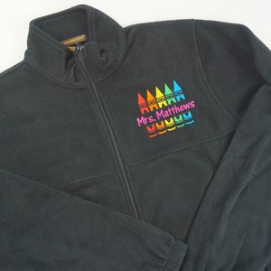 May include: Black fleece zip-up jacket with a colorful crayon design and the name "Mrs. Matthews". The jacket has a high collar and a full-length zipper. The design features red, orange, yellow, green, and blue crayons.