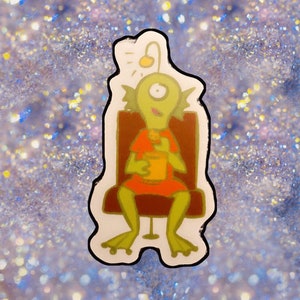 以下が含まれることがあります： 1つの目と頭の上に電球が付いた緑色の漫画の生き物が椅子に座って容器から食べている。生き物は赤いシャツを着ていて、頭の上に緑色のヒレがある。椅子は茶色で白い台座がある。