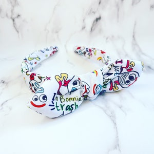 May include: White fabric headband with a colourful cartoon character print. The headband features the text "I'm Bonnie's trash" in a playful font. The design includes various cartoon faces and shapes.