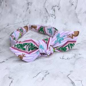 May include: A light purple and white striped headband with a bow. The headband features the text "HONEYDUKES" in green and pink, along with brown and teal frog graphics. The bow is tied in the center.