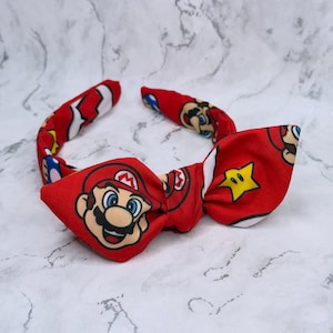 Puede incluir: Pajarita roja con la cara de Mario del videojuego Super Mario Bros. La pajarita tiene un patrón repetido de la cara de Mario, estrellas y otros elementos del juego. La pajarita es de tela.
