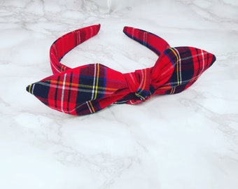 May include: Red and black plaid headband with a large bow. The headband is made of fabric and is a popular accessory for women.