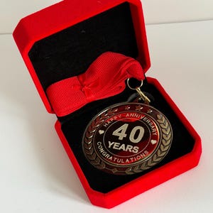 May include: A round medal with a red centre and gold-toned border featuring the text "HAPPY ANNIVERSARY 40 YEARS CONGRATULATIONS" and a red ribbon, presented inside an open, square, red velvet-lined box with a black interior base.