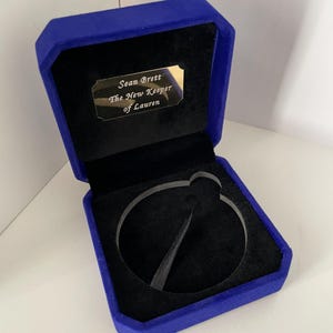 May include: A royal blue velvet jewelry box with a black interior lining. The inside lid features a gold-colored metal plaque engraved with the text: Sean Brett, The New Keeper of Lauren. A circular clear acrylic ring holder sits in the base.