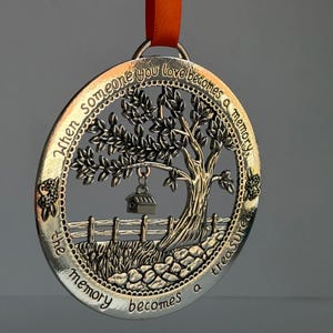 Peut inclure: Un ornement circulaire en métal argenté représentant un arbre en relief, une clôture et un petit nichoir suspendu à une branche. Le texte "When someone you love becomes a memory, the memory becomes a treasure" est gravé sur le bord.
