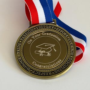 May include: A gold graduation medal with a red, white, and blue ribbon. The medal has the words "On Your Graduation" and "Congratulations" with a graduation cap and diploma design. The medal also has the words "Pride, Passion, Victory, Effort, Courage, Discipline, Respect, Honour" around the edge.