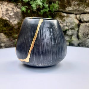 May include: A black ceramic vase with a gold accent. The vase has a textured surface with vertical lines and a rounded shape. A gold line runs diagonally across the vase, adding a decorative touch. The vase is sitting on a white surface.
