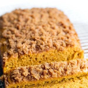 May include: A loaf of pumpkin bread with a streusel topping, sliced and placed on a wire rack. The bread is a light orange color and the streusel is a light brown color.