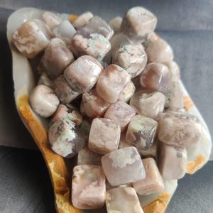 May include: A collection of tumbled stones in shades of pink, cream, and gray, filling a decorative, oblong dish. The stones have a smooth, polished appearance, with some displaying subtle patterns and variations in color. The dish has a light-colored interior and a brown rim.