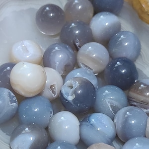 May include: A collection of polished, spherical gemstones in shades of gray, blue, and cream. Some spheres have visible internal crystal formations. The stones are arranged in a natural, light-colored bowl, creating a visually appealing display.