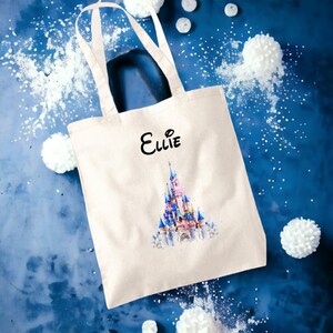 May include: A white canvas tote bag featuring a watercolour illustration of a pink and blue castle. The bag has black handles and the name "Ellie" is printed in black on the front.