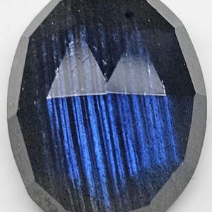 Puede incluir: Gema ovalada con un borde exterior facetado gris oscuro. El interior presenta un llamativo tono azul con estrías verticales y dos formas triangulares en la parte superior. La gema tiene una apariencia texturizada.