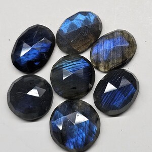 May include: Seven faceted labradorite gemstones in various oval and round shapes. The stones display a range of dark blue, gray, and iridescent flashes of color. The gemstones are arranged on a white surface.