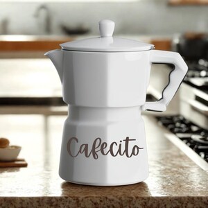 Peut inclure: Cafetière blanche avec couvercle et poignée. Le mot "Cafecito" est écrit en script marron sur le devant. La cafetière est posée sur un comptoir moucheté.