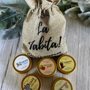May include: A burlap bag with the text "La Tabita! Candle Samples" printed on it. The bag contains four small, gold-colored tins with labels for different scented soy candles: Cayo Coco, Guava, Cubano Rum, and Cafe.