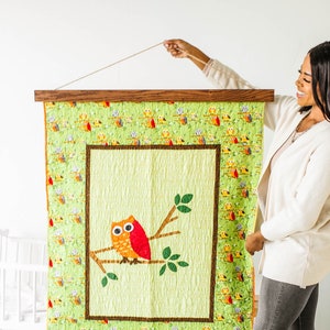 May include: A wooden frame with a green and brown quilt featuring a red and orange owl on a branch. The quilt is hanging on a wall with a string.