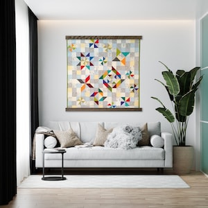 May include: A colorful quilt with a geometric pattern of stars and squares, hanging on a white wall in a living room setting. The quilt is framed with a wooden frame and is displayed above a white sofa with pillows.