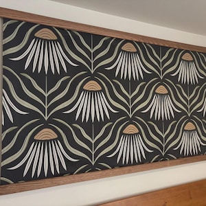 May include: A framed wall hanging with a repeating floral pattern. The design features white, tan, and green flowers with long, curved leaves on a black background. The frame is made of light brown wood.